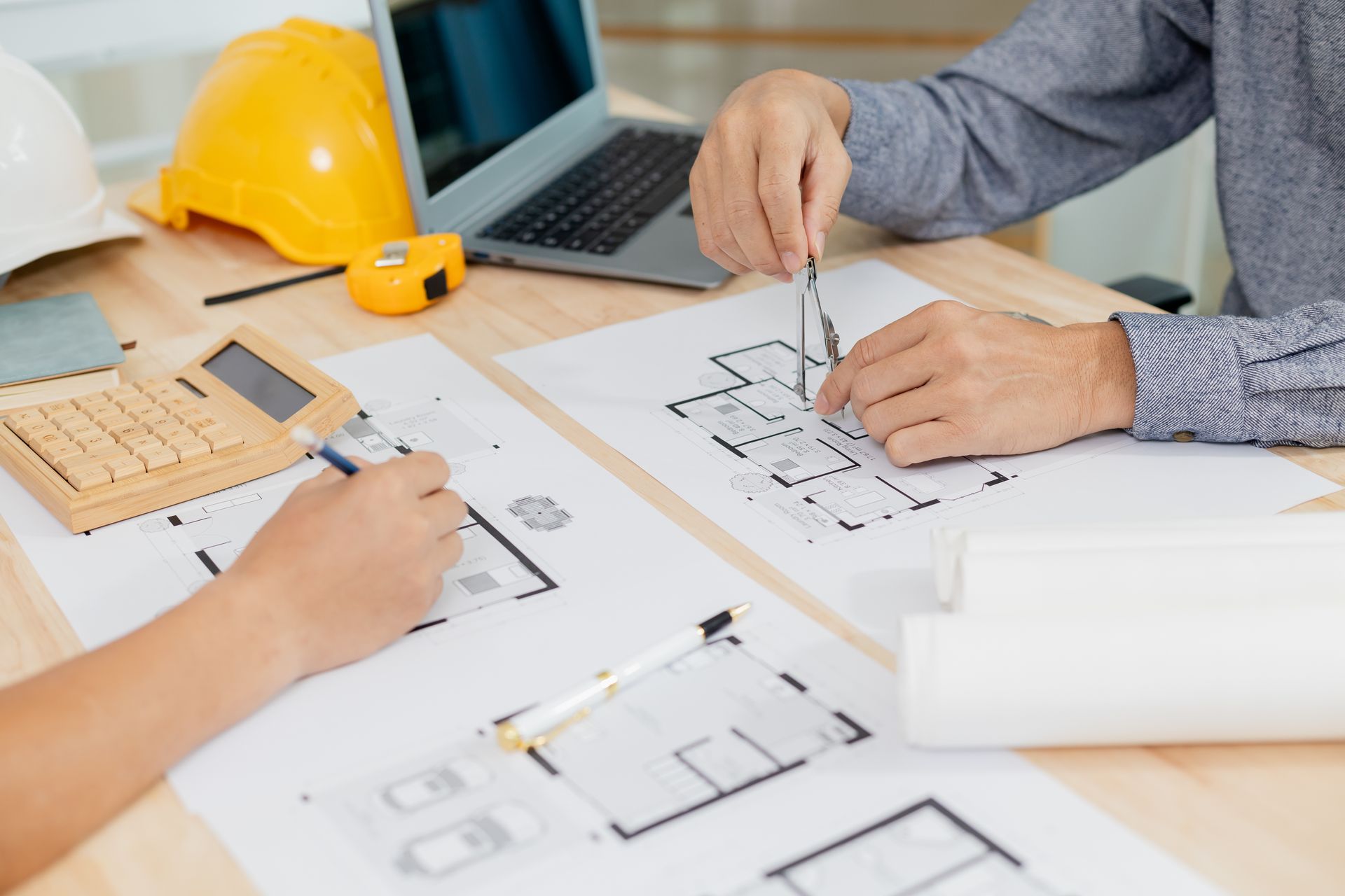 Hands drafting house plans at a desk with laptop, ruler, and yellow hard hat