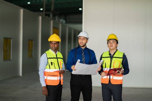Three construction workers in hard hats and safety vests reviewing blueprints in an unfinished building.