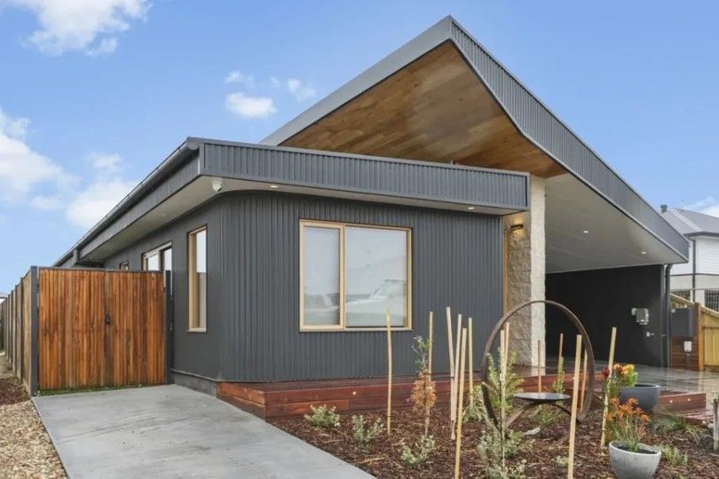 Modern gray house with gabled roof, wood accents, driveway, and landscaped front yard under a blue sky Modern gray house with gabled roof, wood accents, driveway, and landscaped front yard under a blue sky