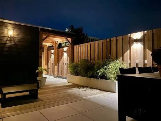 Nighttime rooftop patio with wooden pergola, planters, lounge chairs, and warm wall lights Nighttime rooftop patio with wooden pergola, planters, lounge chairs, and warm wall lights
