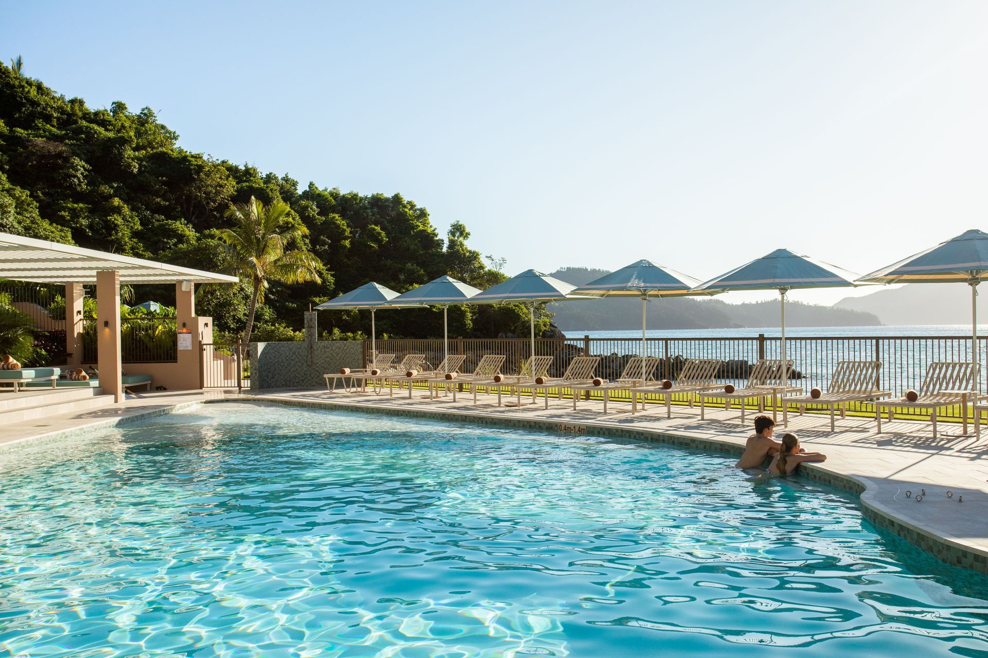 Sunny resort pool with lounge chairs, umbrellas, and people relaxing by the water