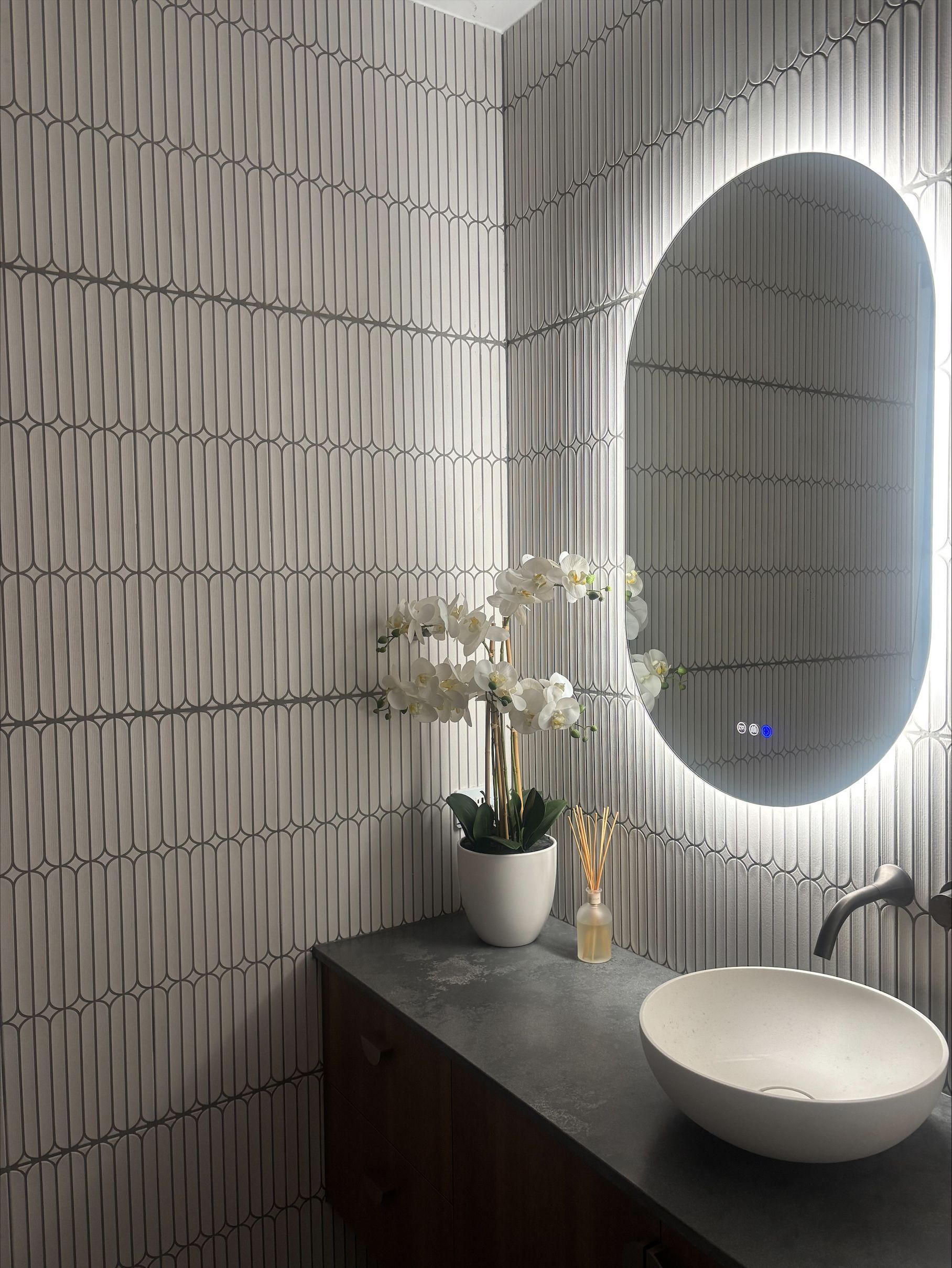 Modern bathroom vanity with oval backlit mirror, white vessel sink, dark countertop, and white flowers