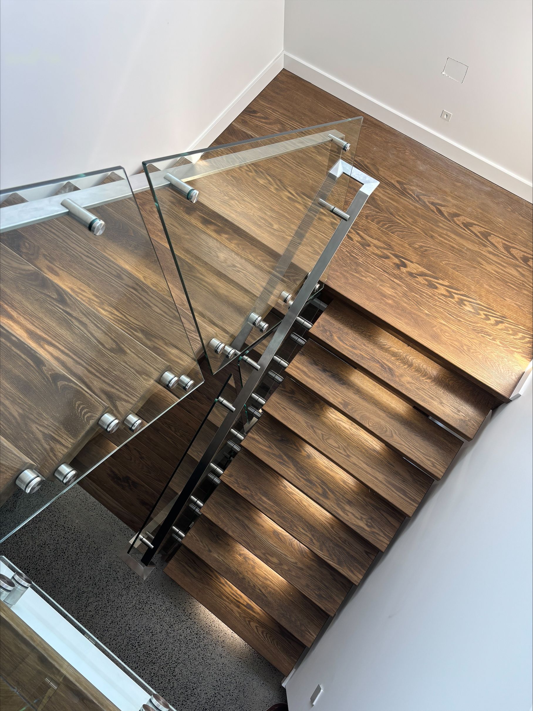 Top-down view of a wooden staircase with a glass railing and white walls.
