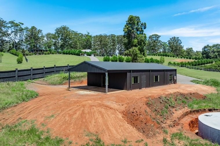 Small dark house under construction on a dirt lot, with a fenced green hillside in the background.