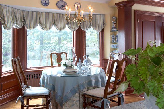 Dining room with round table, wooden chairs, chandelier, and large windows with a green leafy plant.