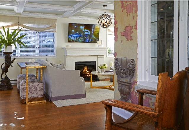 Bright living room with fireplace, wall-mounted TV, gray sofa, wooden chairs, and large windows