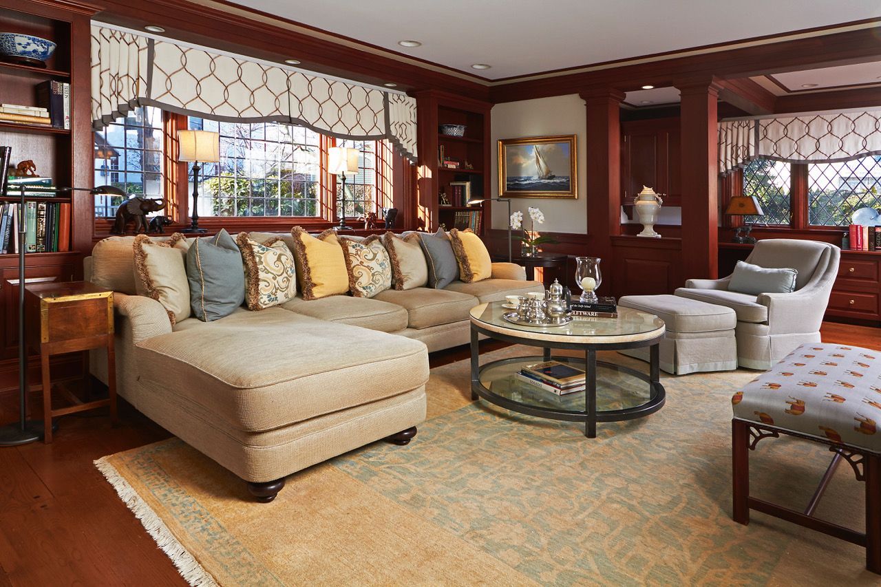 Warm living room with beige sectional, glass coffee table, red walls, and patterned chairs by large windows