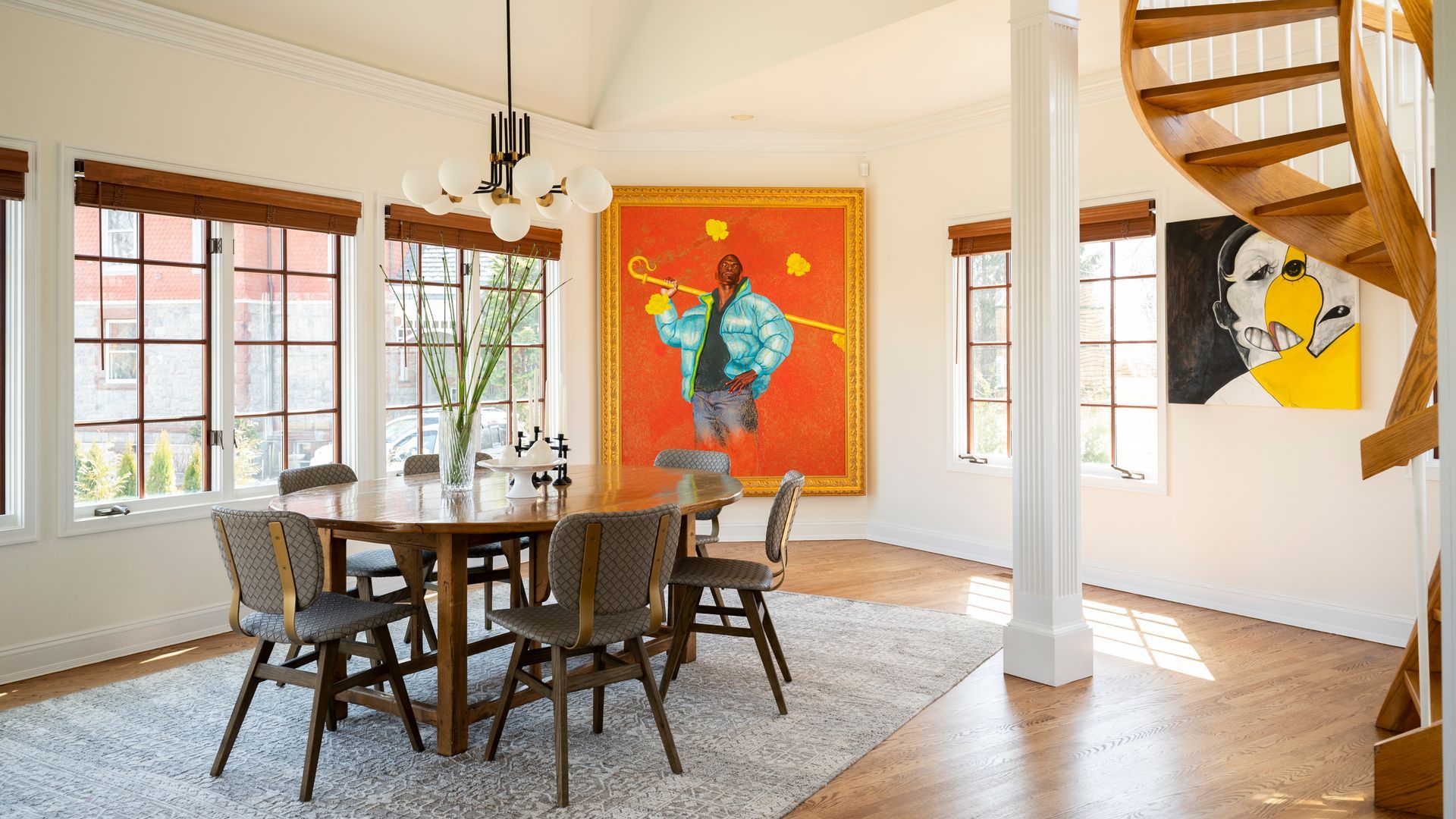 Bright dining room with a wooden table, patterned rug, colorful wall art, and a spiral staircase.