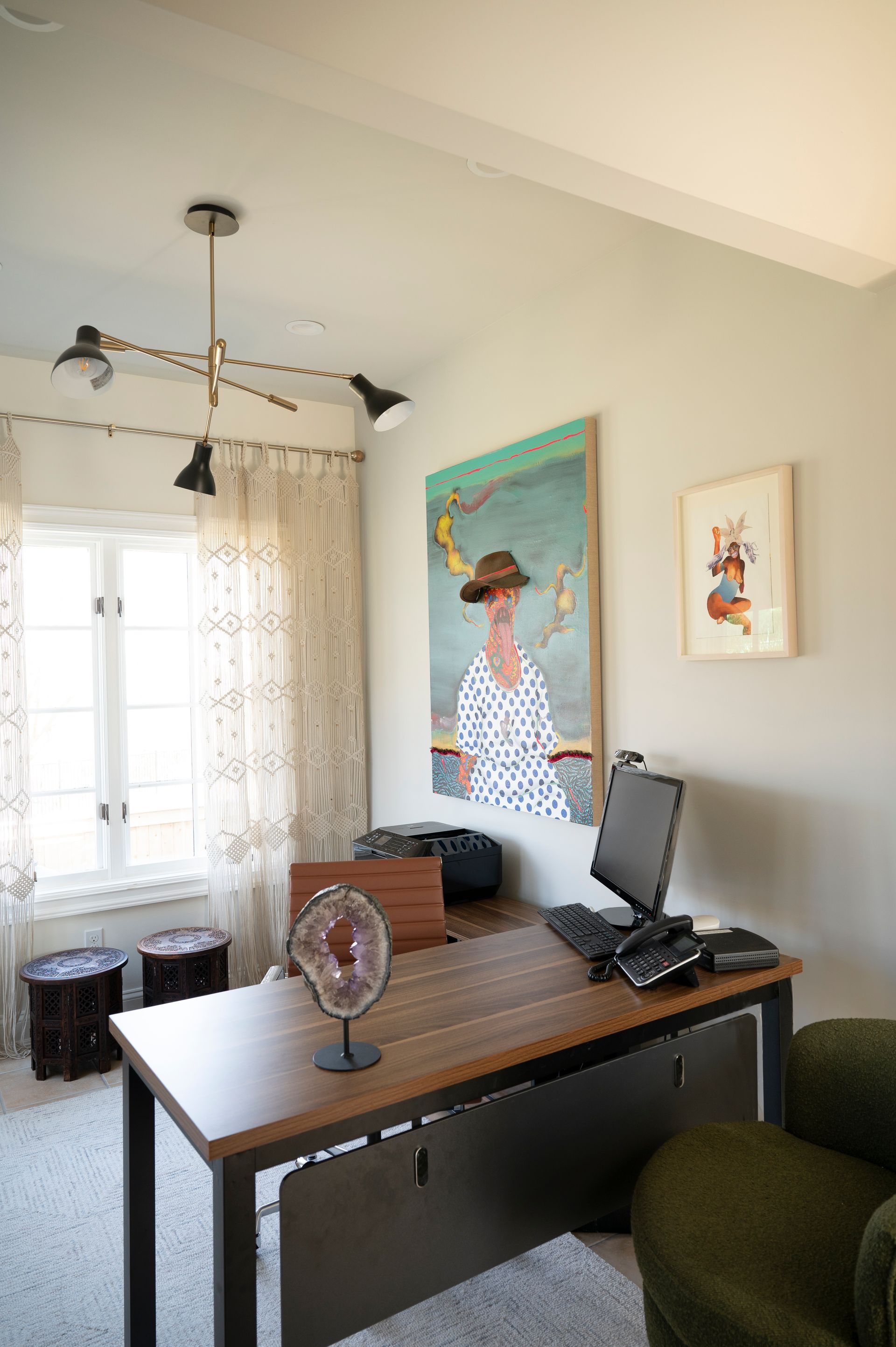 Small home office with a wooden desk, green chair, window, and wall art in a bright room.