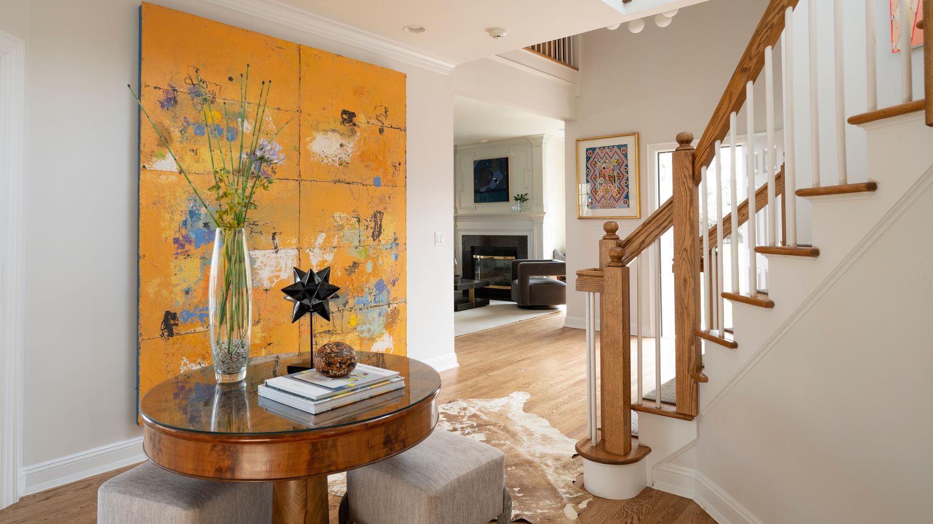 Bright entryway with orange wall art, round table, and staircase with white spindles