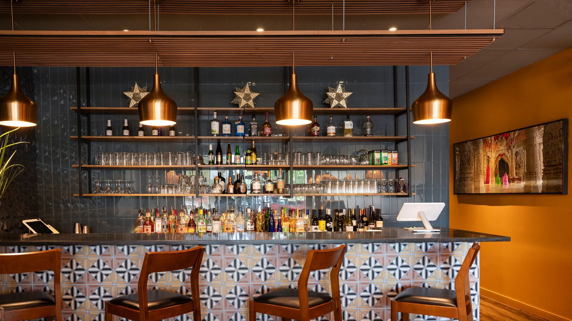 Warm modern bar interior with wooden counter, hanging pendant lights, shelves of bottles, and stools.