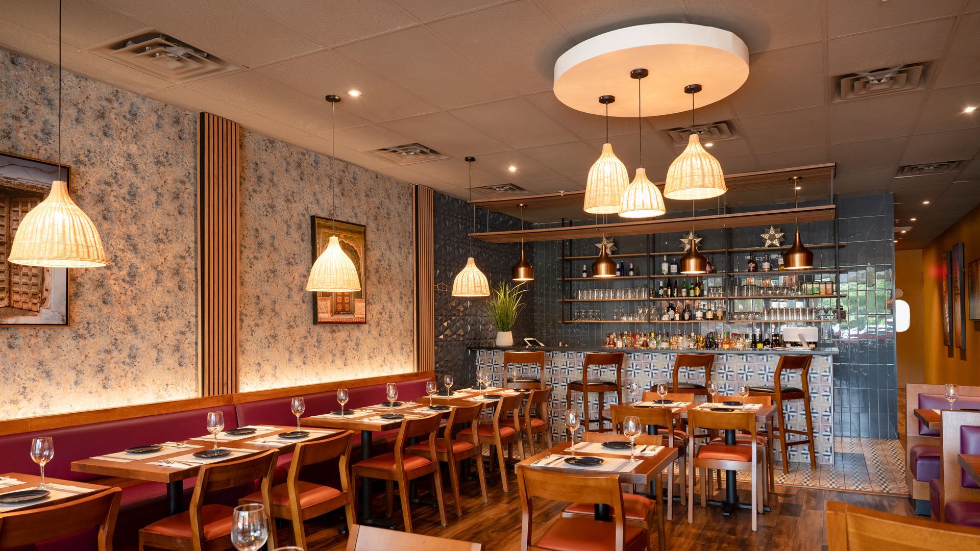Warm restaurant dining room with wooden tables, pendant lights, patterned walls, and a bar in back