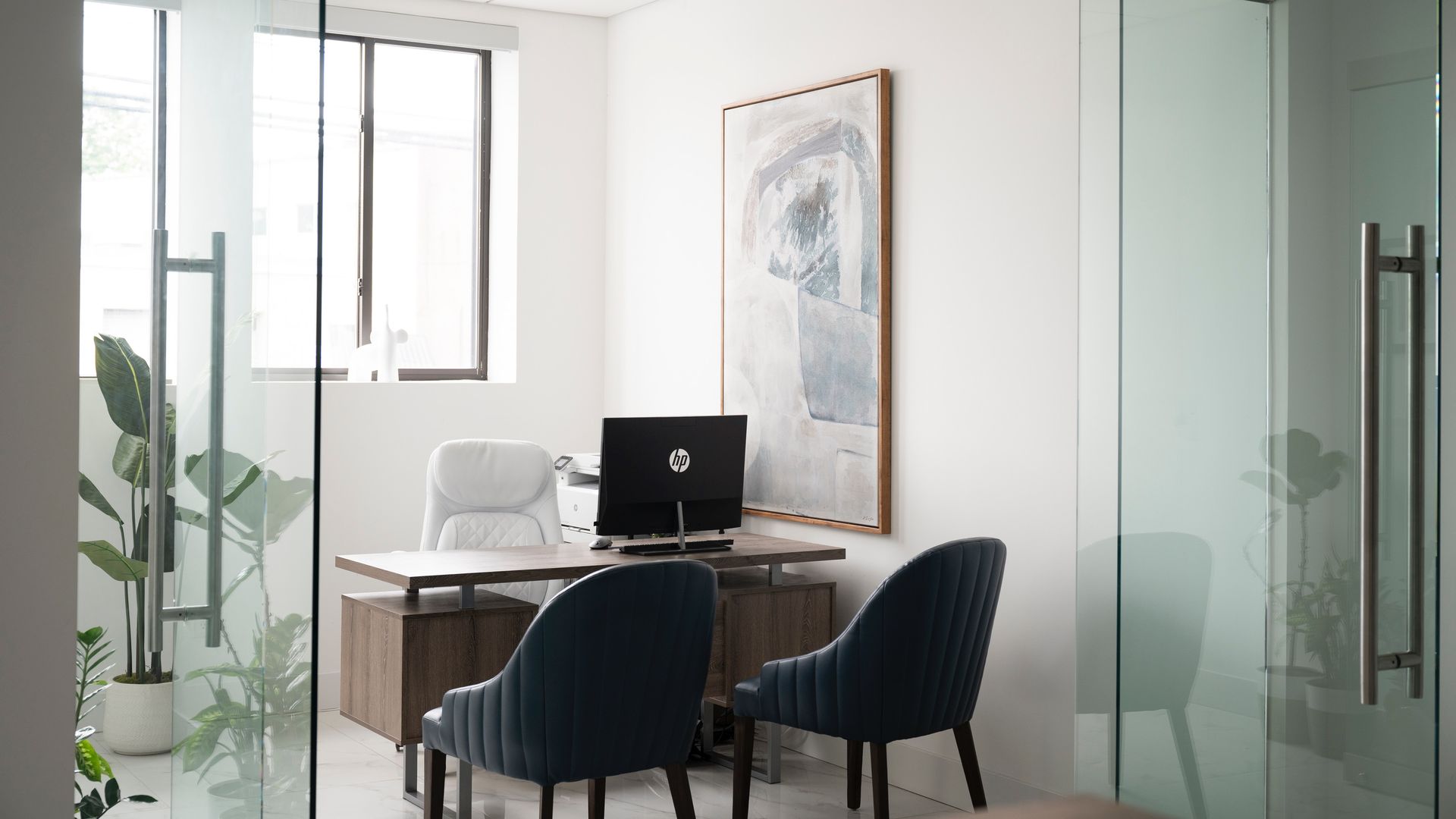 Modern office meeting nook with desk, laptop, and two dark chairs beside glass walls