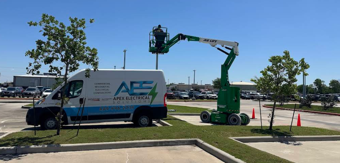 Green boom lift beside a white utility van in a sunny parking lot, with trees and traffic cones nearby