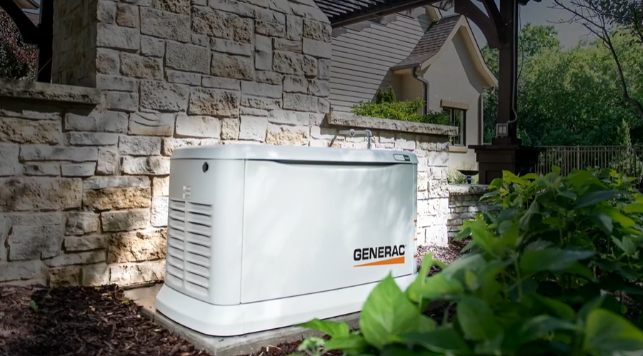 White standby generator next to a stone wall in a yard, with a house in the background.