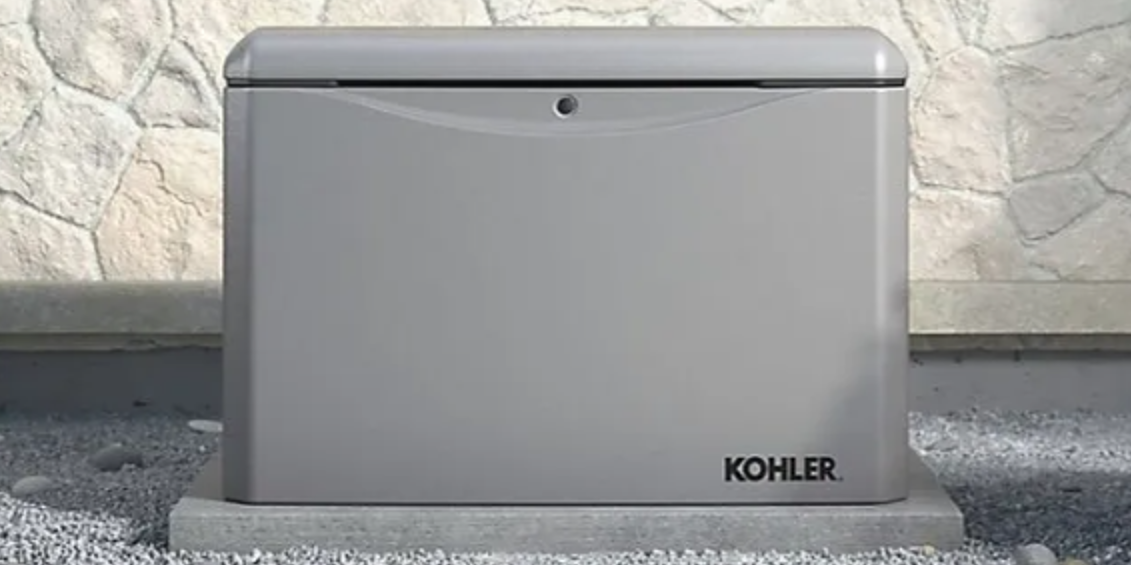 A gray Kohler generator box on a concrete pad with a stone wall backdrop.