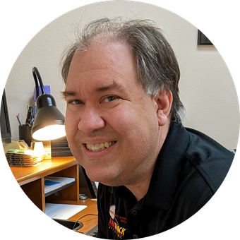 Man smiles, sitting at a desk with a lamp. Black shirt.