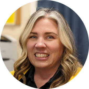 Woman with blonde and brown wavy hair smiles, wearing a yellow jacket and black shirt, indoors.