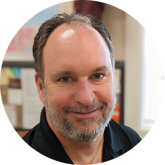 Man with a graying beard, smiling, wearing a black shirt, in an office setting.