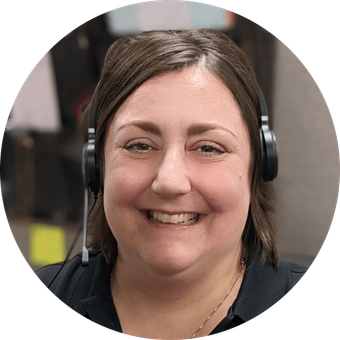 Woman wearing a headset, smiling at the camera. She appears to be in an office setting.