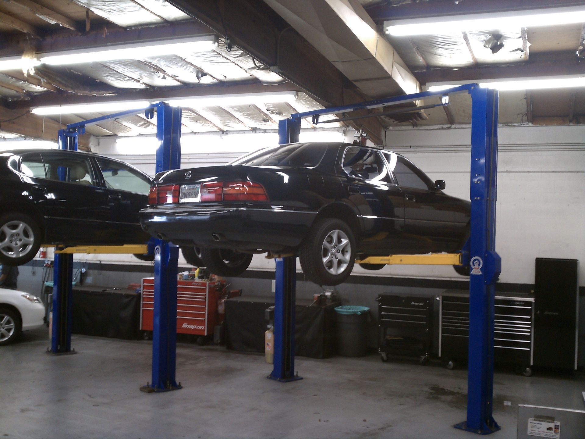 Two cars on blue hydraulic lifts in an auto repair shop with fluorescent lighting.