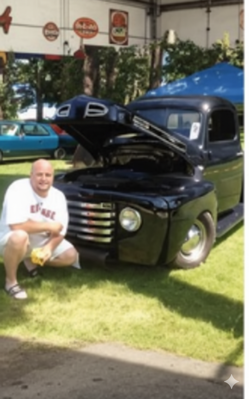 Man kneeling beside a black vintage pickup truck with its hood open at a car show outdoors