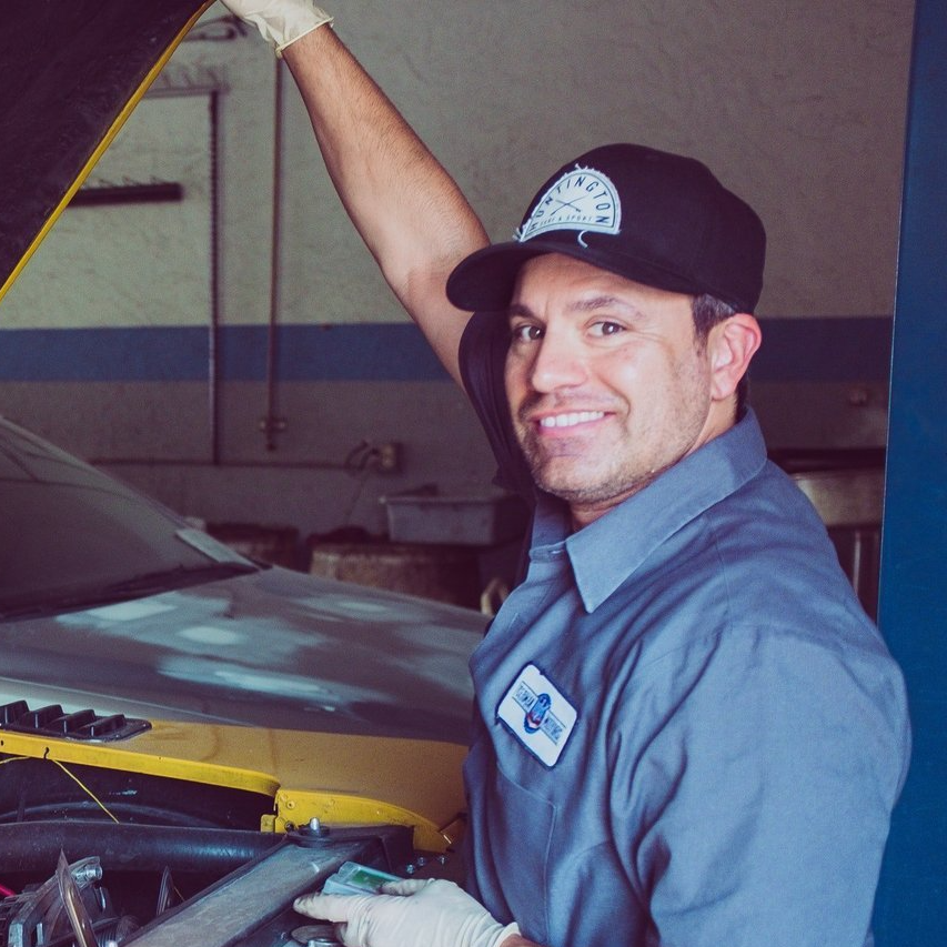 Mechanic smiling in a garage, holding a car hood open beside a yellow vehicle.