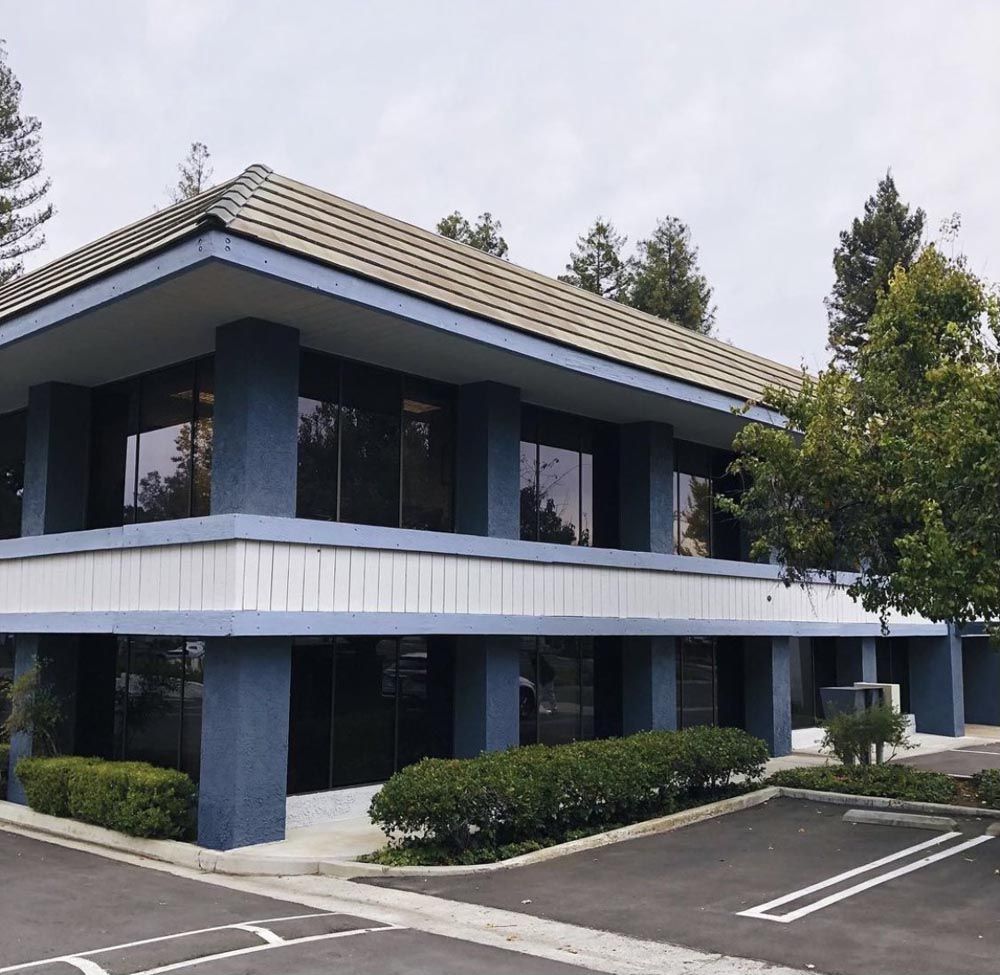 Two-story blue and white commercial building with large windows and tiled roof, trees and parking lot in foreground.