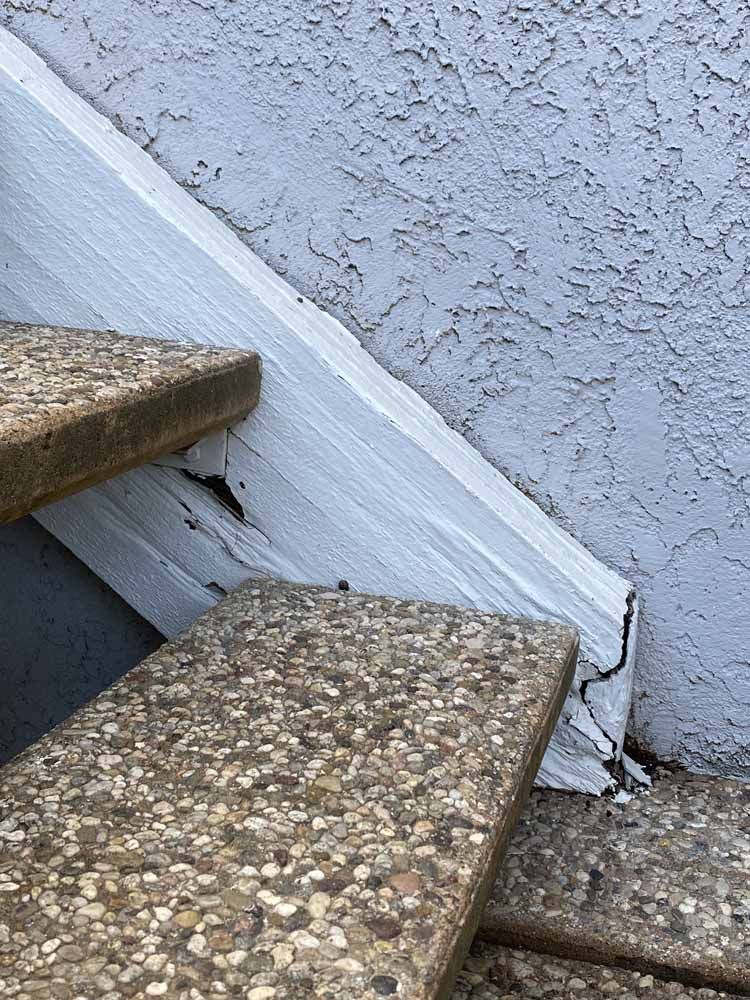 Concrete steps next to a weathered white wooden beam against a textured gray wall.