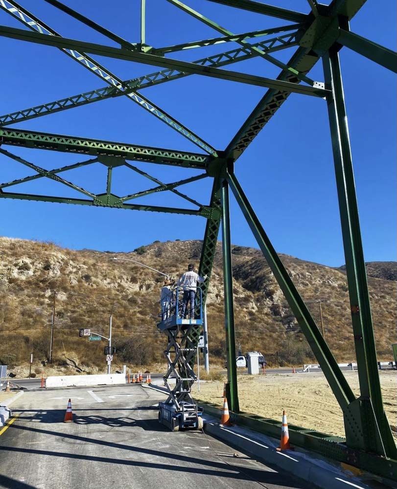Person on lift painting green bridge framework against a clear blue sky, mountain backdrop.