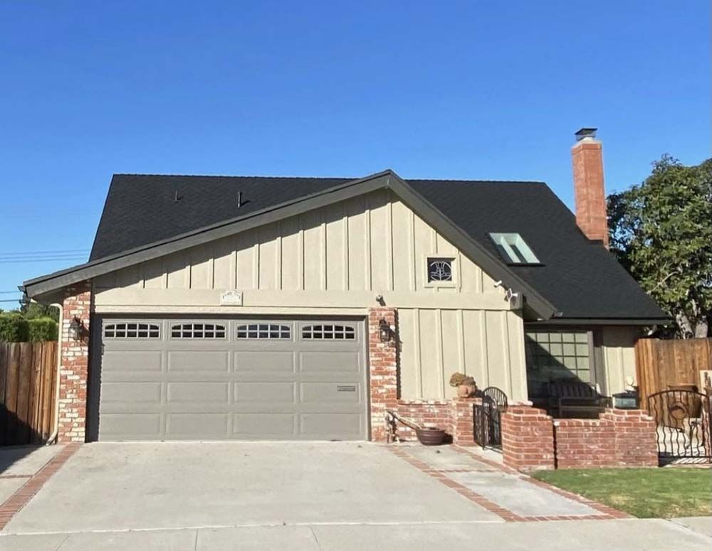 House with gray garage door, tan siding, brick accents, and a black roof against a blue sky.