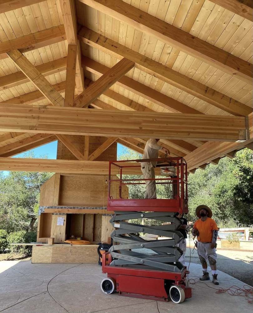 Workers on a lift under a wooden roof structure, building in an outdoor setting.