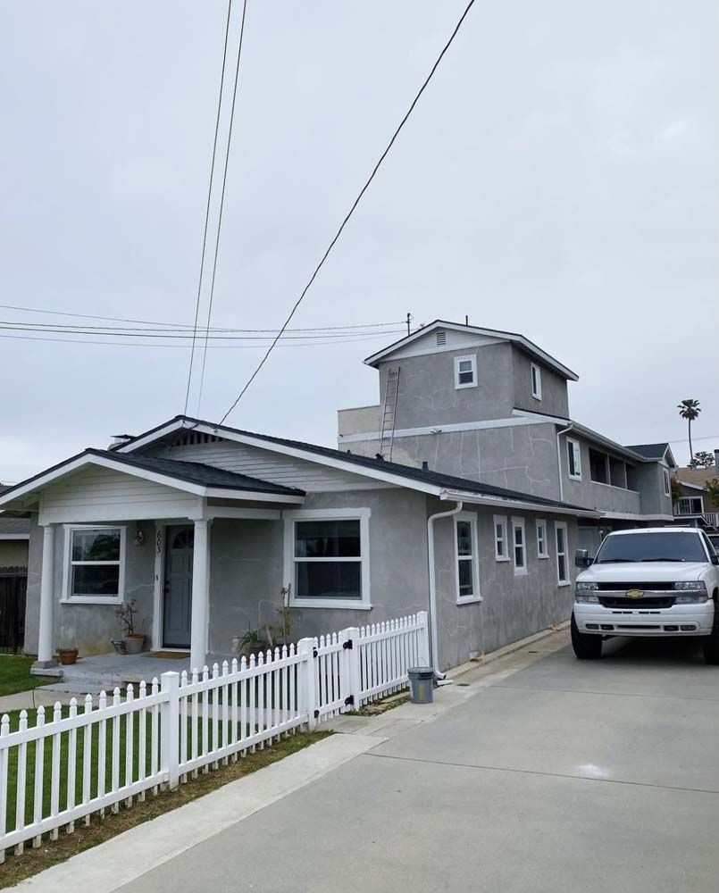 A gray house with a white picket fence and a white truck parked in the driveway. Cloudy sky.
