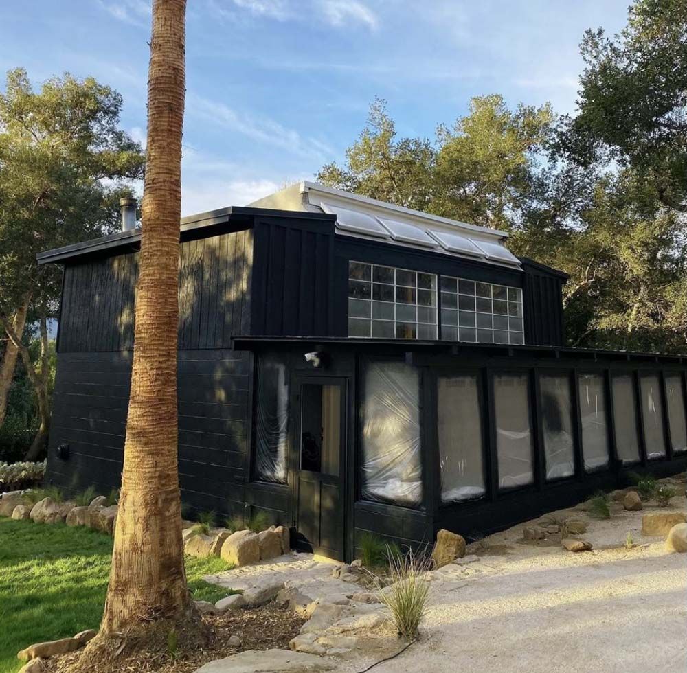 Black building with glass windows and a transparent roof panel, surrounded by trees and a paved pathway.