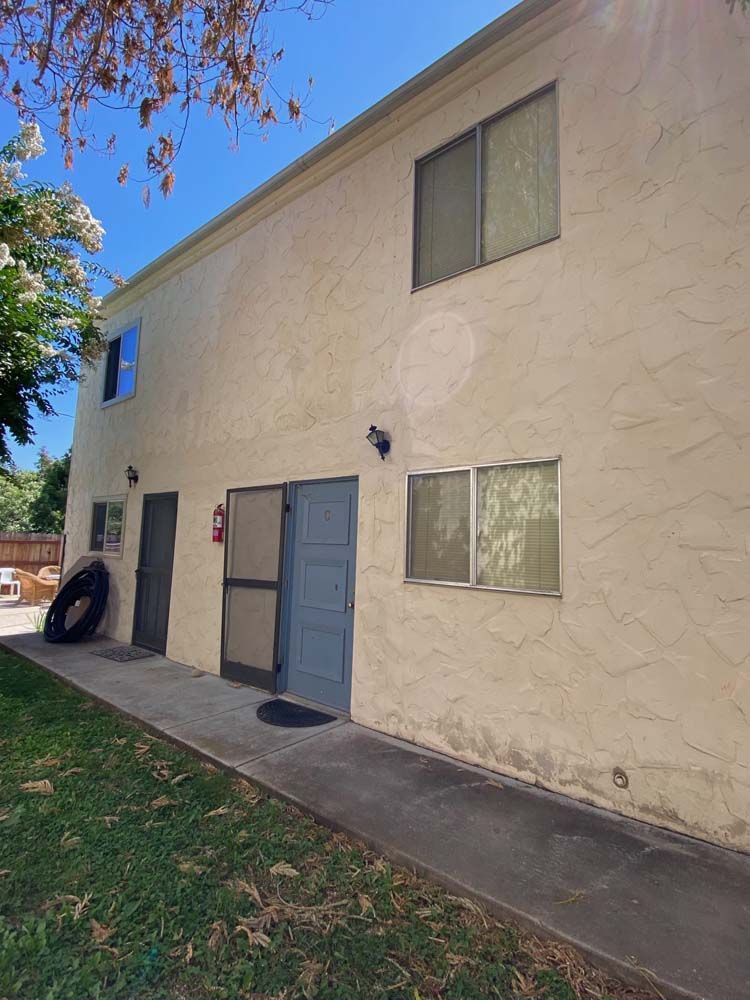 Two-story beige stucco building with windows and doors, on a concrete path, under a blue sky.