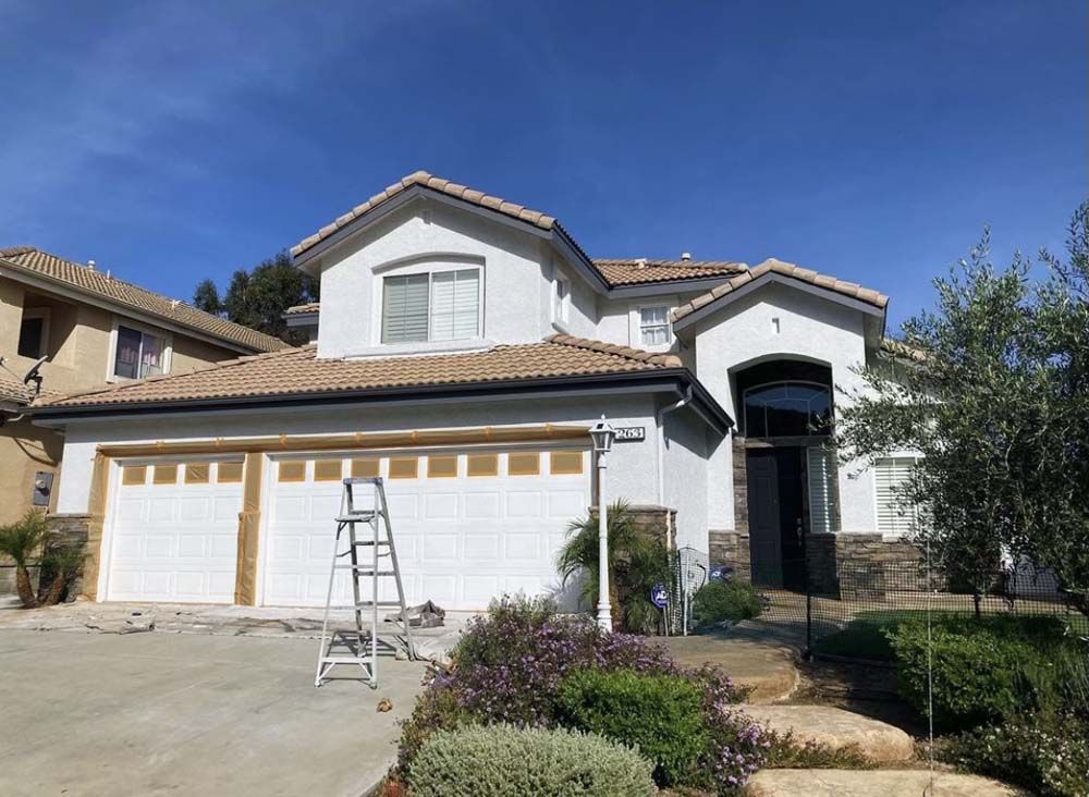 Two-story white house with brown roof and garage doors, ladder in front, landscaping, and a blue sky.