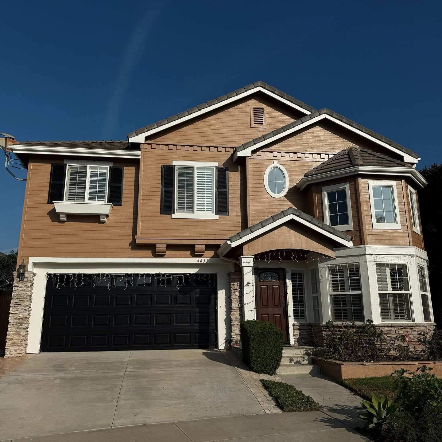 Suburban two-story house with tan siding, stone accents, black garage door, and front porch under a blue sky