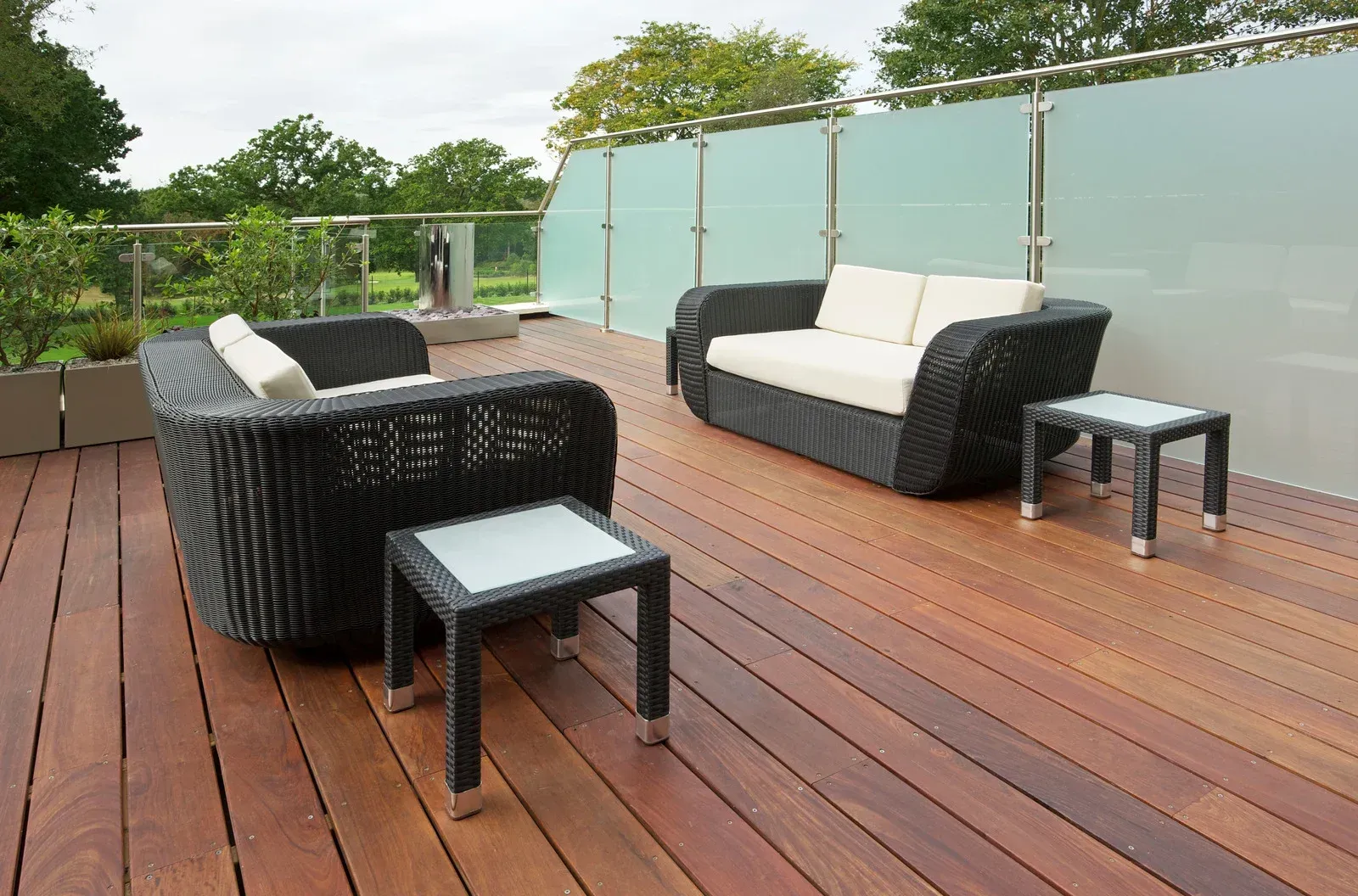 Outdoor seating area on a wooden deck with dark wicker furniture and glass tables, overlooking trees.