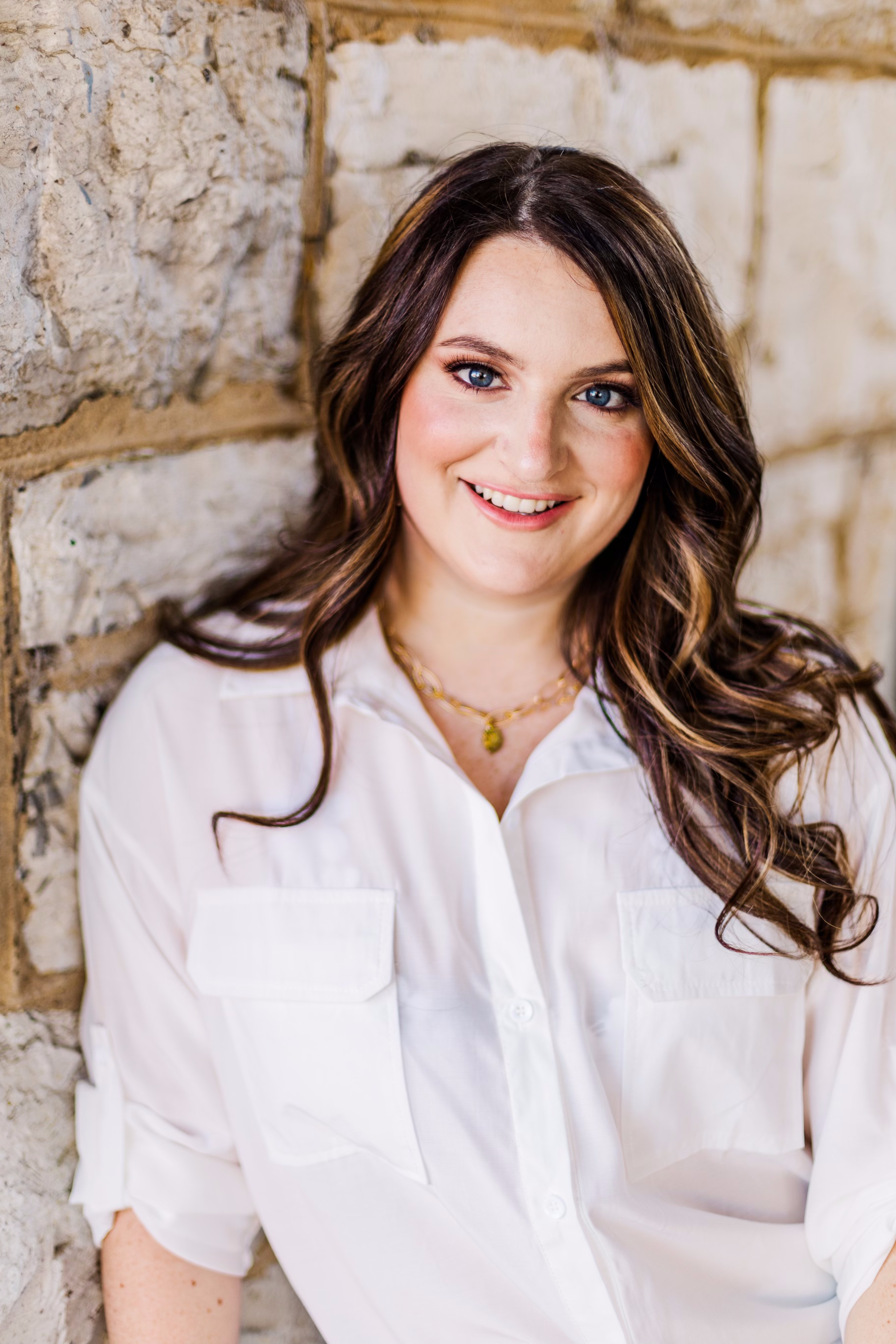 A woman in a white shirt is leaning against a stone wall and smiling.