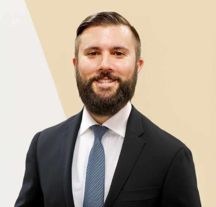 Smiling man in a dark suit and blue tie against a beige background