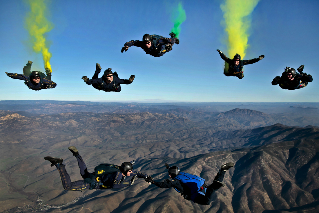 Skydivers in black suits with colored smoke trails descend over a mountain landscape.