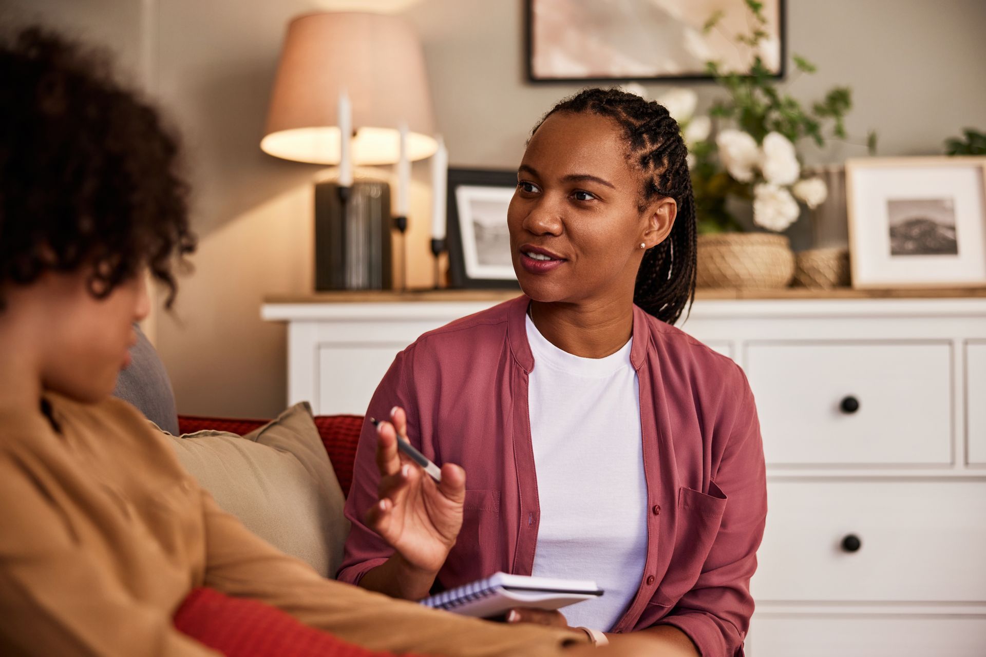 A person in a pink shirt speaks and gestures while holding a notebook, sitting with another person in a living room.