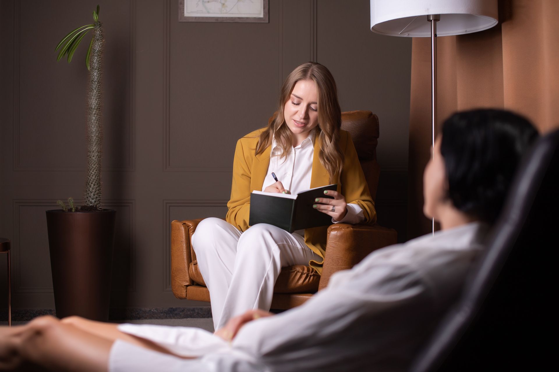 A therapist in a yellow blazer sits in a chair, taking notes while a patient lies on a couch in a calm office.