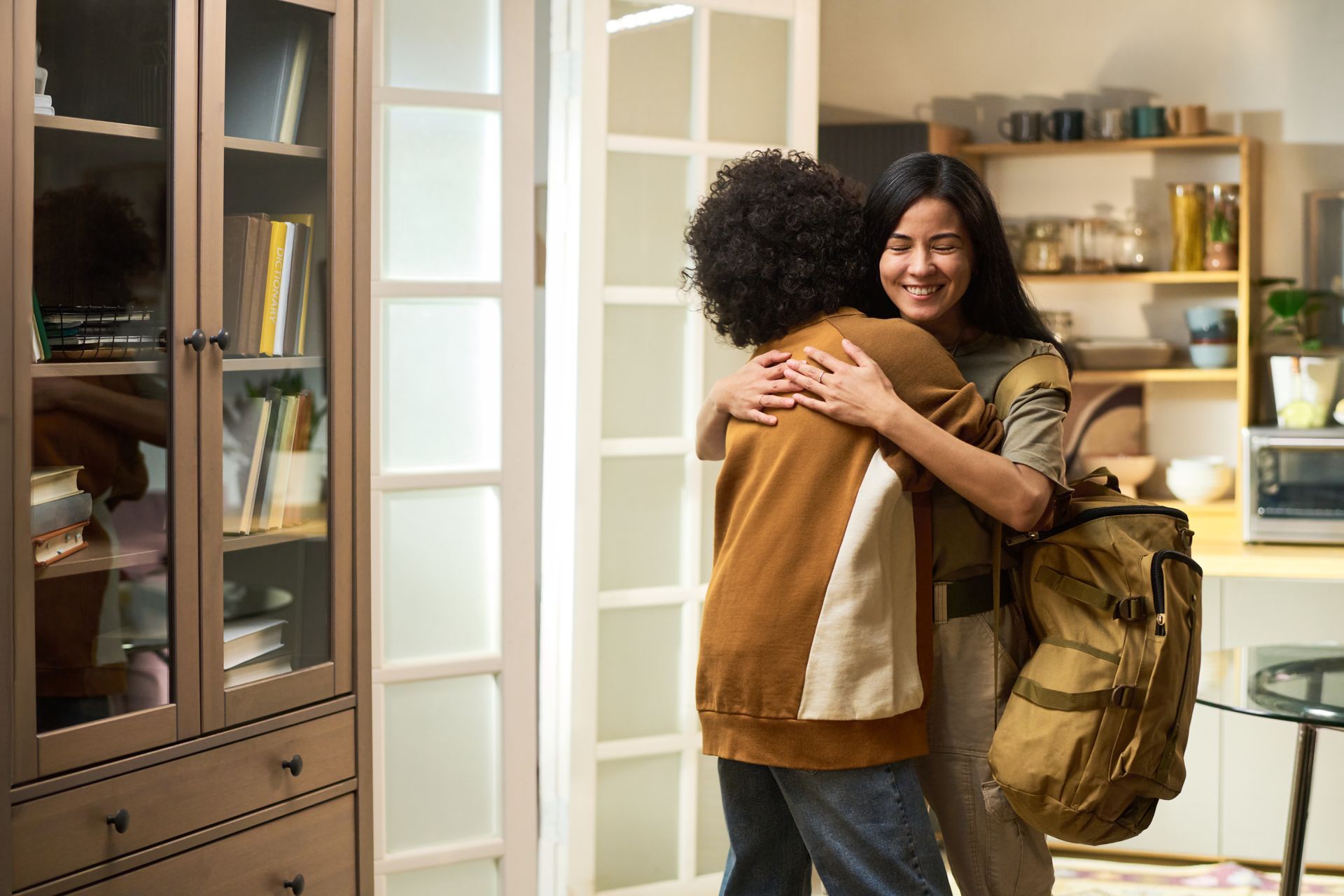 Two people embrace warmly in a home setting. One person wears a tan backpack, and a wooden bookshelf is visible nearby.