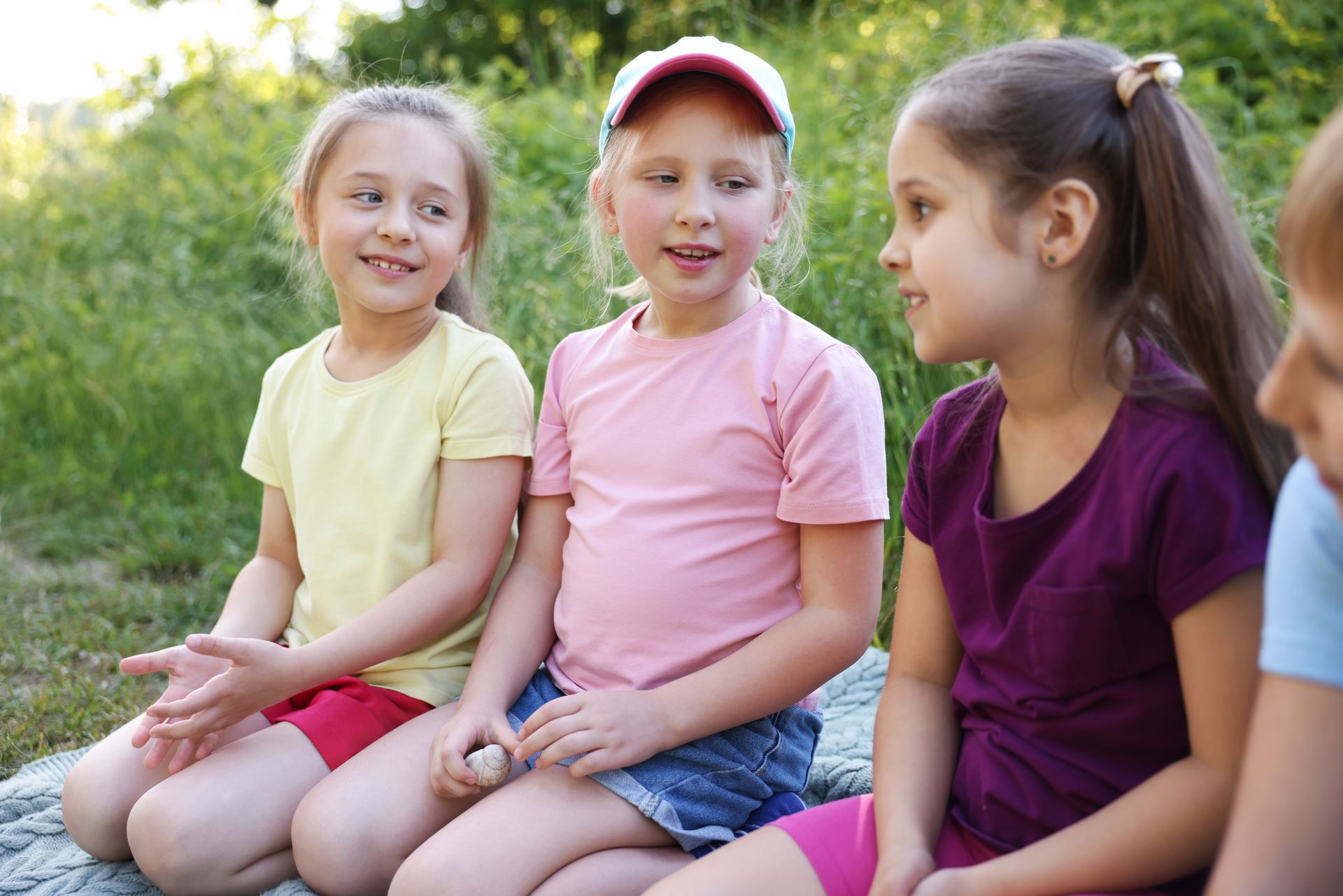 Three children sit together outdoors in a grassy area, smiling and talking to one another.