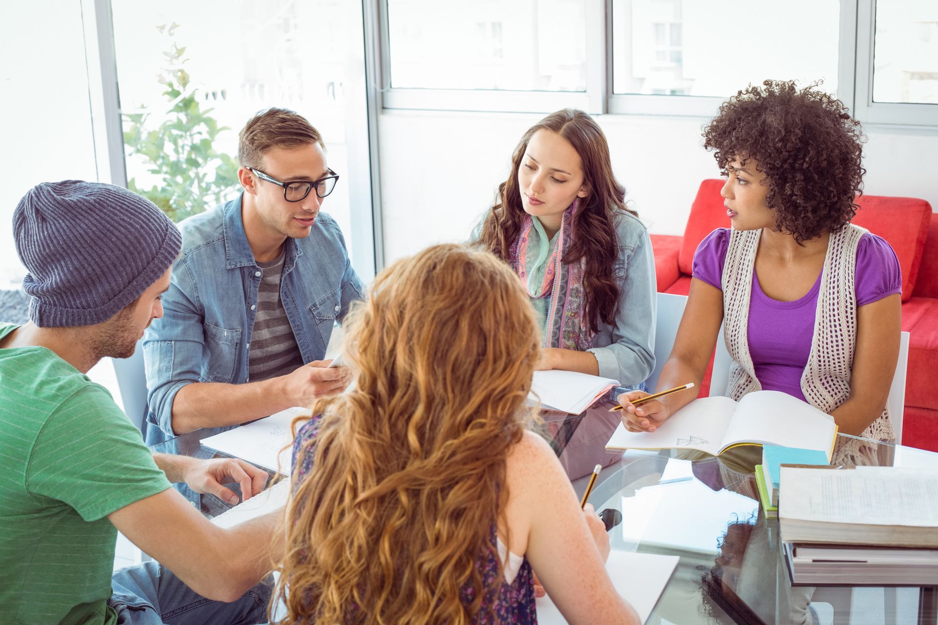 Four people sit around a table studying, writing in notebooks and looking at each other during a collaborative discussion.