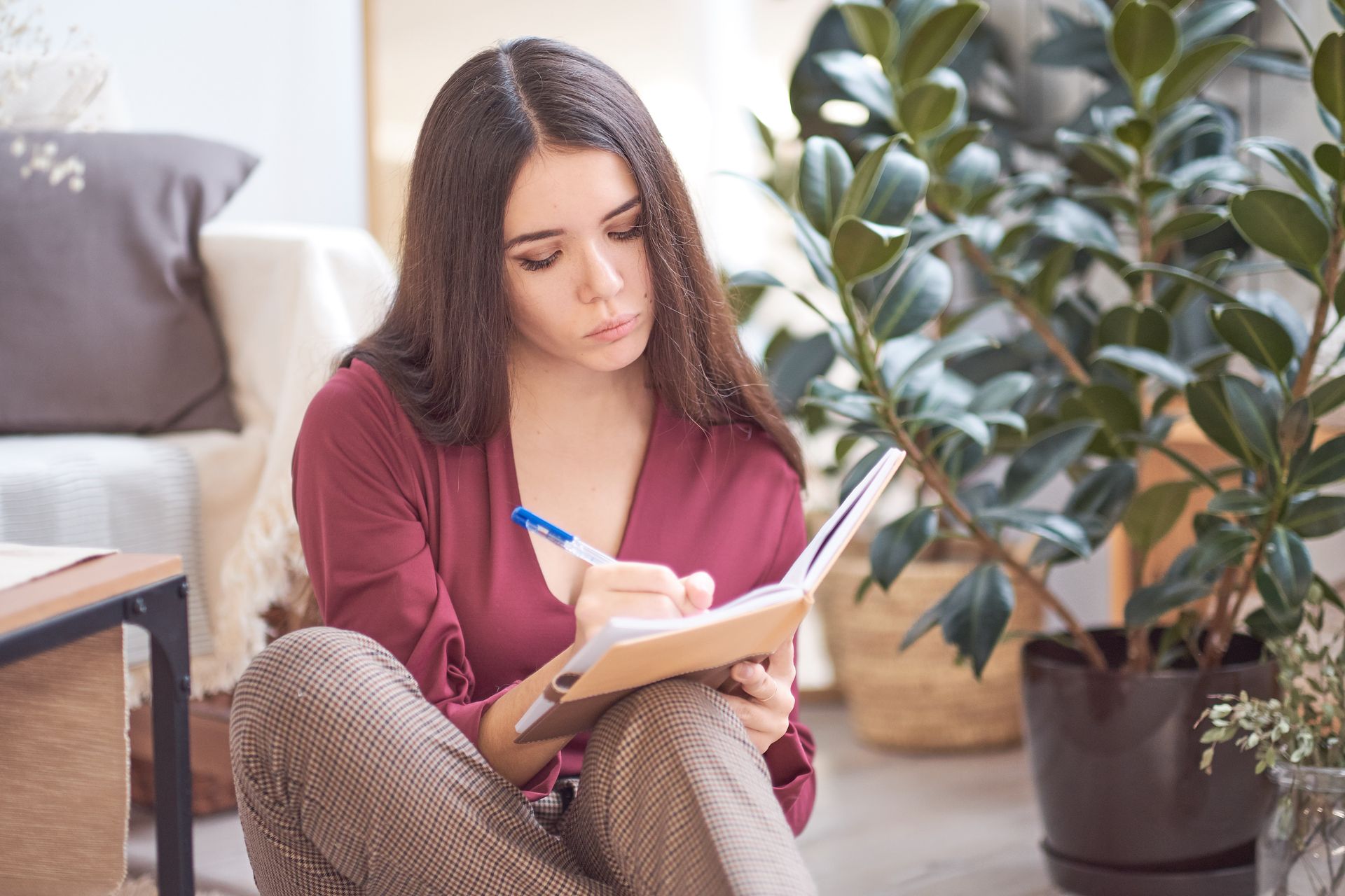 A person in a maroon shirt sits on the floor writing in a notebook, with a leafy houseplant and part of a couch nearby.