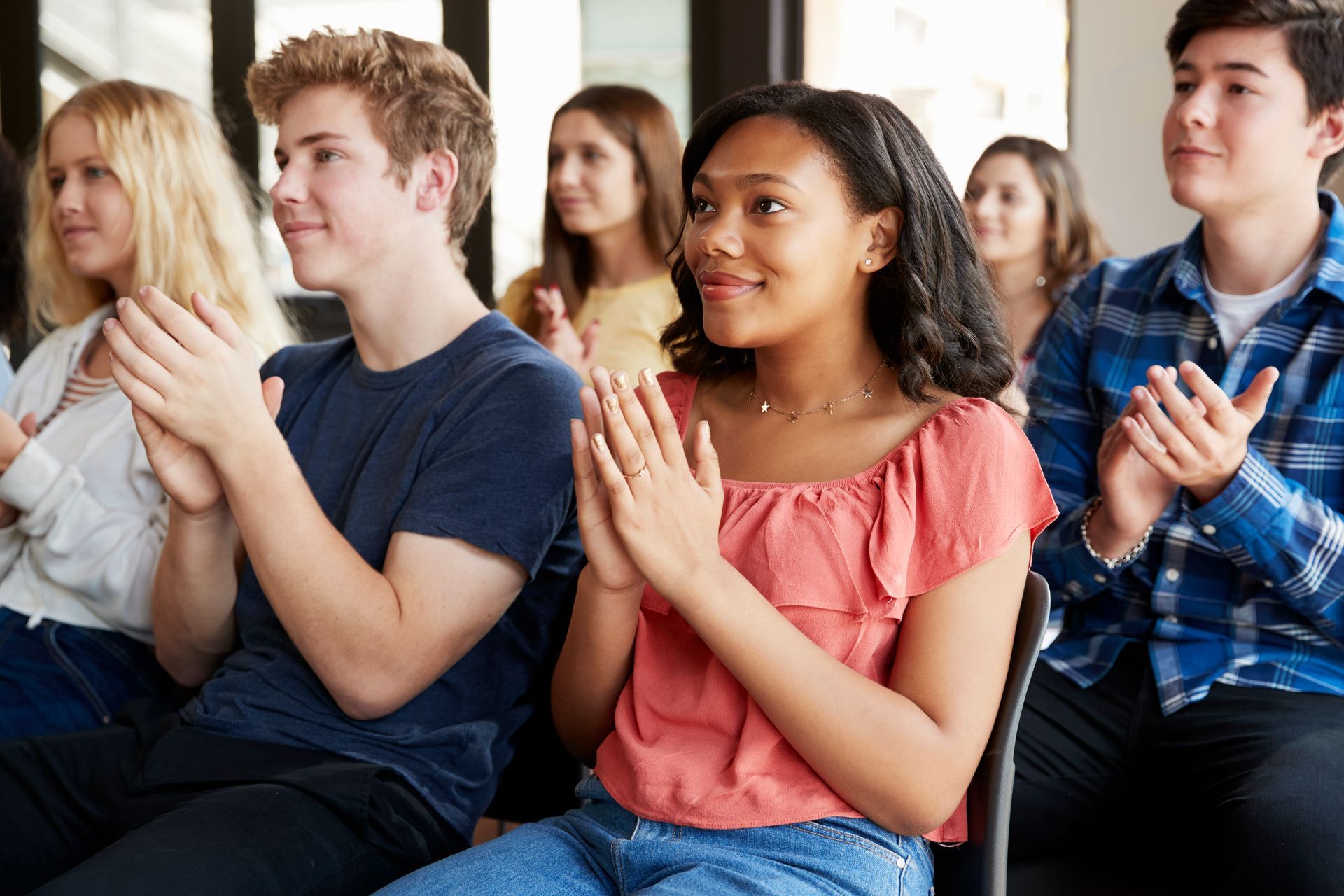 A group of people sitting in chairs and clapping, appearing to be an audience at a presentation or event.