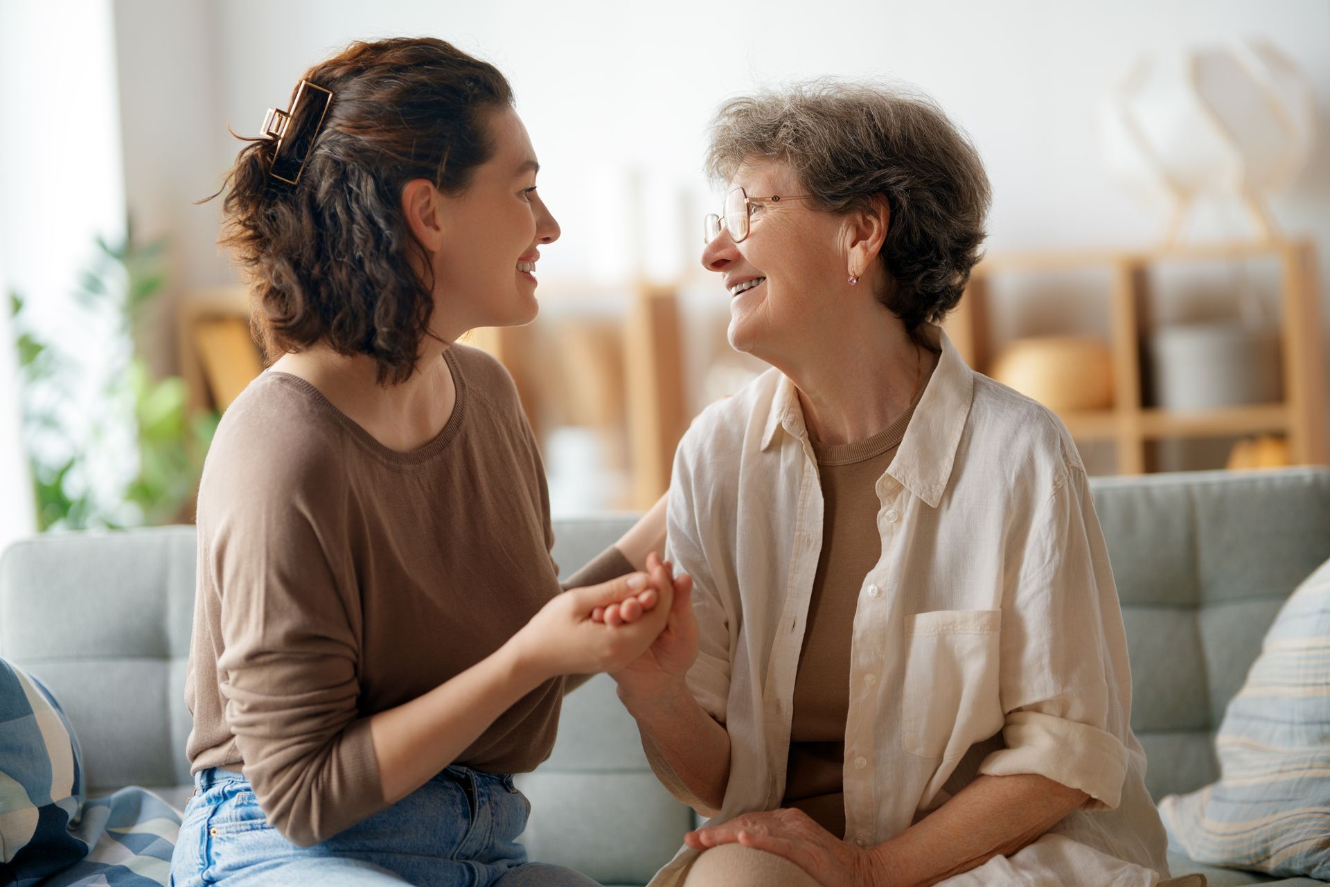 A young person and an older person sit on a sofa, holding hands and smiling at each other in a brightly lit room.