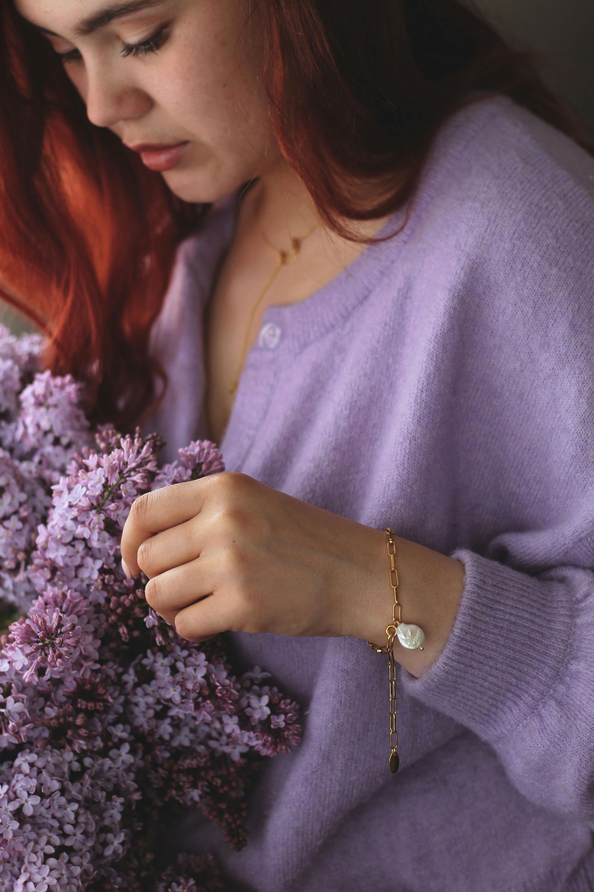 Woman in a lavender sweater holding purple flowers, looking down with a gold bracelet on her wrist